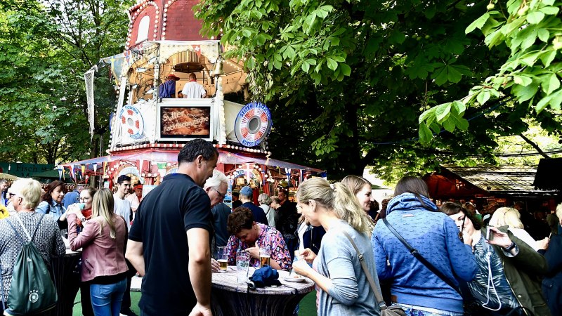 Menschen genießen Speisen und Getränke vor einem rot-weißen Imbissstand auf einem Markt, umgeben von grünen Bäumen., © michael schwartz photographie Menschen genießen Speisen und Getränke vor einem rot-weißen Imbissstand auf einem Markt, umgeben von grünen Bäumen., © michael schwartz photographie