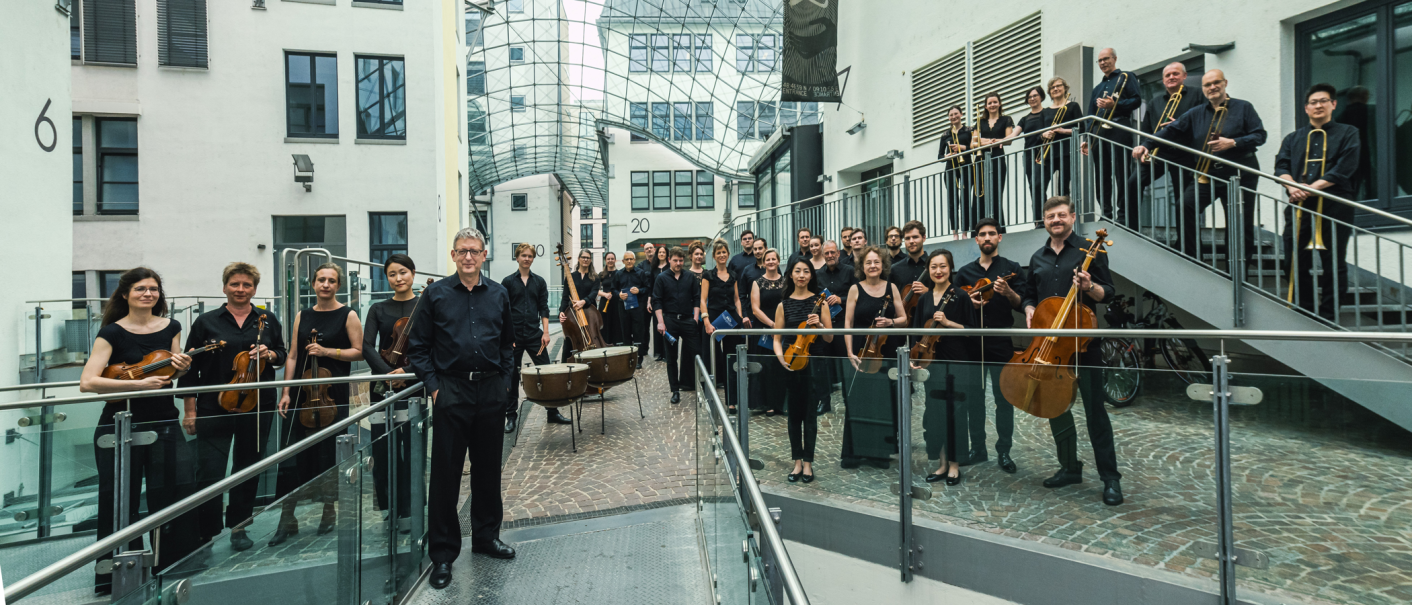 Die Gaechinger Cantorey posiert in einem modernen Innenhof. Die Musiker tragen schwarze Kleidung und halten verschiedene Instrumente., &copy; Martin F&ouml;rster