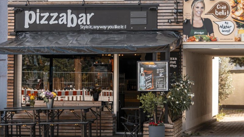 Au&szlig;enansicht einer Pizzabar mit Terrasse, Blumen und einem Werbeplakat. Ein Mann sitzt an einem Tisch. Die Markise ist schwarz., &copy; Stuttgart Marketing GmbH, Sarah Schmid