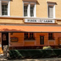 Ein Restaurant namens Fischlabor mit orangener Markise und Sitzgelegenheiten im Freien. Das Gebäude hat gelbe Ziegelwände und weiße Fensterrahmen., © SMG, Sarah Schmid Ein Restaurant namens Fischlabor mit orangener Markise und Sitzgelegenheiten im Freien. Das Gebäude hat gelbe Ziegelwände und weiße Fensterrahmen., © SMG, Sarah Schmid