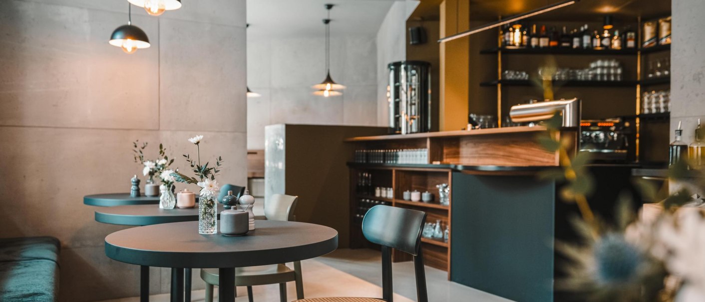Modernes Café mit eleganter Einrichtung, runden Tischen und Blumenvasen. Dezente Beleuchtung und eine gut ausgestattete Bar im Hintergrund., © Lupin, Stuttgart