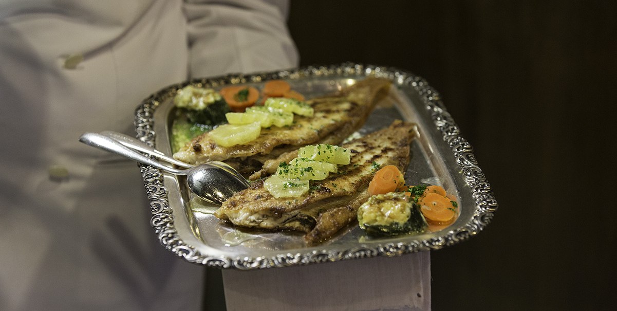 Ein silbernes Tablett mit gegrilltem Fisch, garniert mit Zitrone und Gemüse, wird von einem Kellner in weißer Uniform serviert., © Hotel Royal