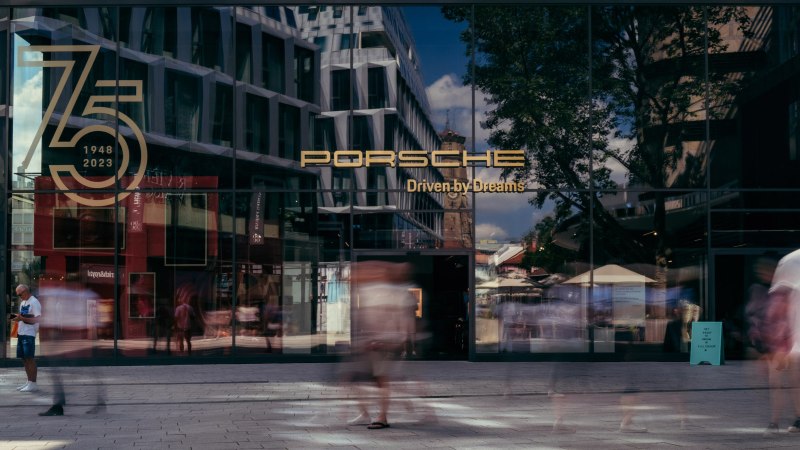 Glass façade of the Porsche Brand Store with anniversary logo '75 years 1948-2023'. People walk past, buildings and trees are reflected in the glass., © Porsche AG