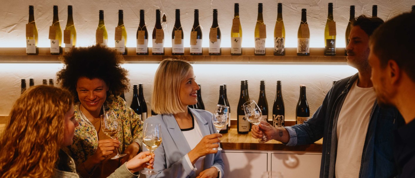 Menschen in einer Vinothek stoßen mit Weingläsern an. Im Hintergrund sind Weinflaschen auf Regalen zu sehen., © SMG, Alwin Maigler Menschen in einer Vinothek stoßen mit Weingläsern an. Im Hintergrund sind Weinflaschen auf Regalen zu sehen., © SMG, Alwin Maigler