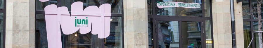 Ein Caf&eacute; mit gro&szlig;en Fenstern und dem Schriftzug 'juni essentials'. Davor stehen Fahrr&auml;der und Tische mit St&uuml;hlen., &copy; SMG, Sarah Schmid