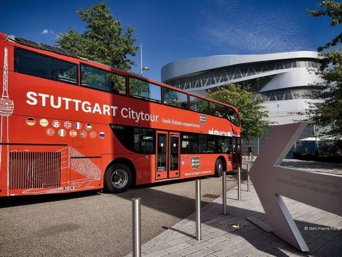 Ein roter Doppeldeckerbus der Stuttgart Citytour steht vor einem modernen, geschwungenen Gebäude. Bäume und blauer Himmel im Hintergrund., © SMG, Pierre Polak Ein roter Doppeldeckerbus der Stuttgart Citytour steht vor einem modernen, geschwungenen Gebäude. Bäume und blauer Himmel im Hintergrund., © SMG, Pierre Polak