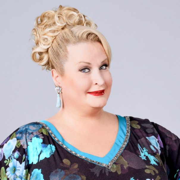 A woman with blonde curls and a floral dress smiles at the camera. She is wearing eye-catching earrings and red lipstick., &copy; Renitenztheater Stuttgart e.V.