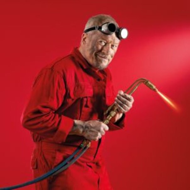 An older man in a red overall is holding a welding machine. He is wearing safety goggles on his forehead. The background is also red., © Altes Schauspielhaus und Komödie im Marquardt e.V. An older man in a red overall is holding a welding machine. He is wearing safety goggles on his forehead. The background is also red., © Altes Schauspielhaus und Komödie im Marquardt e.V.