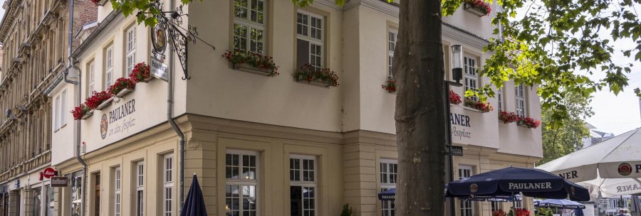 Eckgebäude mit Paulaner-Restaurant, dekoriert mit roten Blumen. Vor dem Gebäude stehen Sonnenschirme und Tische. Bäume spenden Schatten., © SMG, Sarah Schmid