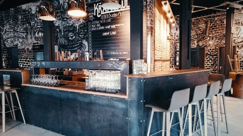 Moderne Bar mit industriellem Design, hohen Stühlen und Wandkunst. Gläser stehen auf der Theke, im Hintergrund eine Tafel mit Getränken., © Kraftpaule