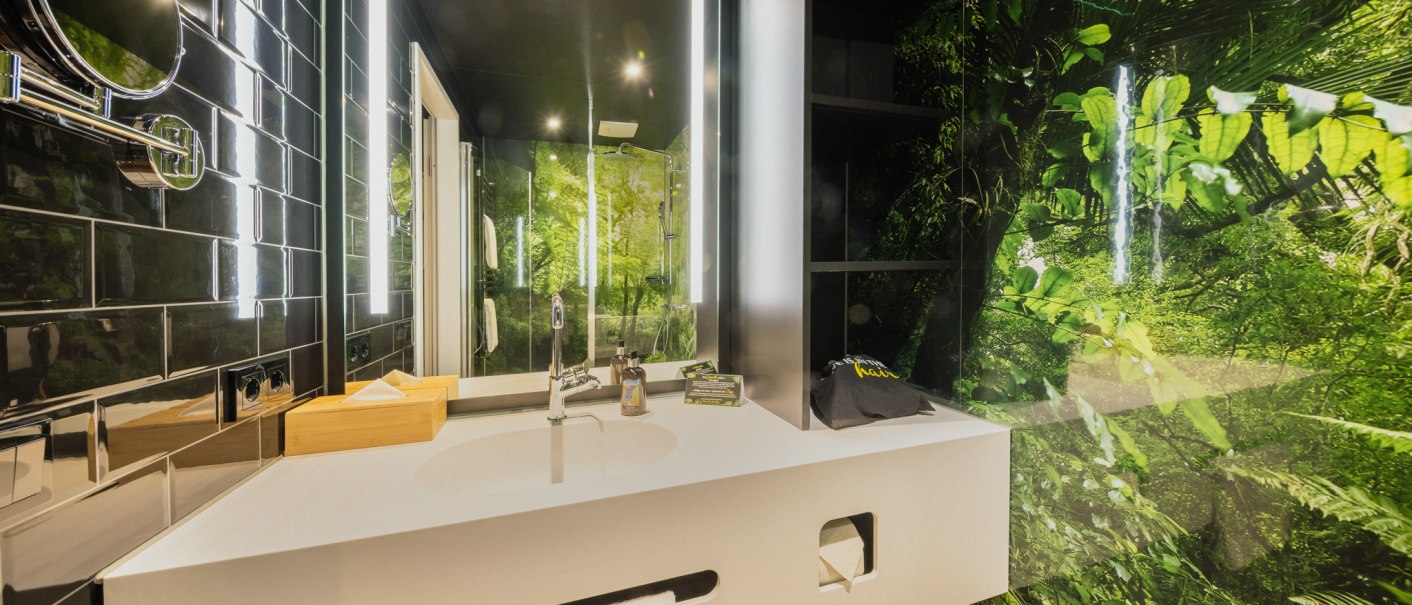 Modern bathroom with white washbasin, mirror and jungle motif on the wall. Black tiles and bright lighting complete the design., © Rioca Stuttgart Posto 4 GmbH Modern bathroom with white washbasin, mirror and jungle motif on the wall. Black tiles and bright lighting complete the design., © Rioca Stuttgart Posto 4 GmbH