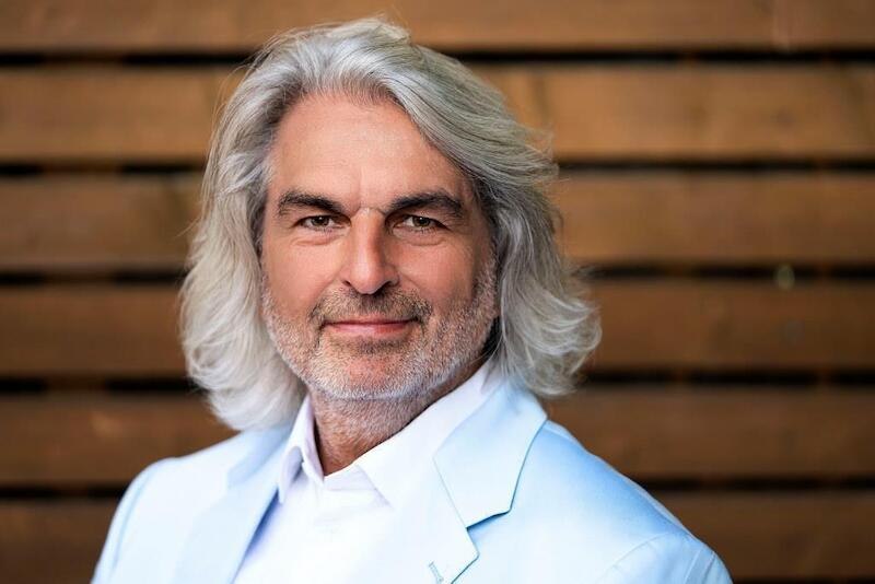 Ein Mann mit grauem Haar und Bart trägt einen weißen Anzug. Er steht vor einem Holzhintergrund und lächelt leicht., © Rosenau Kultur e.V. Ein Mann mit grauem Haar und Bart trägt einen weißen Anzug. Er steht vor einem Holzhintergrund und lächelt leicht., © Rosenau Kultur e.V.