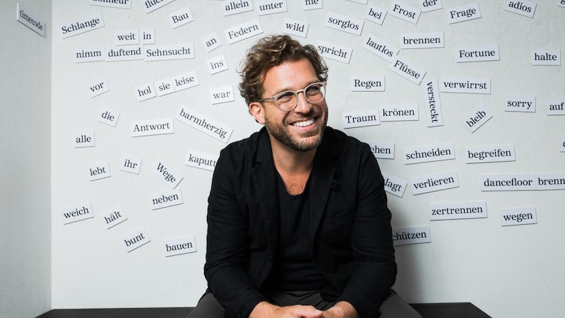 Ein Mann mit Brille sitzt lächelnd vor einer Wand, die mit verschiedenen deutschen Wörtern beklebt ist., © Internationale Hugo-Wolf-Akademie Ein Mann mit Brille sitzt lächelnd vor einer Wand, die mit verschiedenen deutschen Wörtern beklebt ist., © Internationale Hugo-Wolf-Akademie