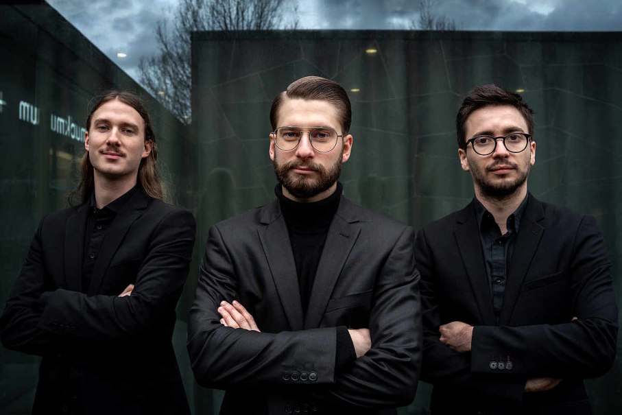 Drei Männer in schwarzen Anzügen stehen mit verschränkten Armen vor einer Glaswand. Der Himmel ist bewölkt., © JH-Photography