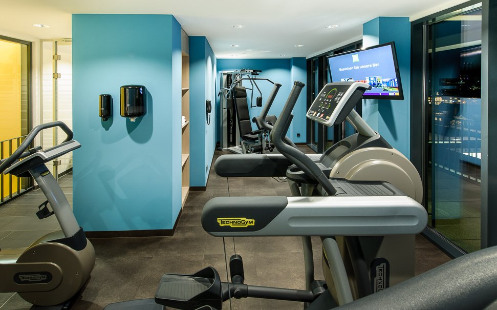 Moderner Fitnessraum mit blauen Wänden, ausgestattet mit Laufbändern, Fahrrädern und einem Bildschirm. Große Fenster bieten Blick nach draußen., © TOMAS Moderner Fitnessraum mit blauen Wänden, ausgestattet mit Laufbändern, Fahrrädern und einem Bildschirm. Große Fenster bieten Blick nach draußen., © TOMAS