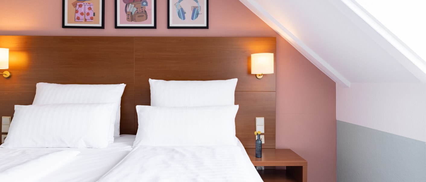 A modern hotel room with two single beds, white sheets, and pillows. Three pictures hang above the headboard. The walls are painted pink and gray., &copy; Novum  Hospitality