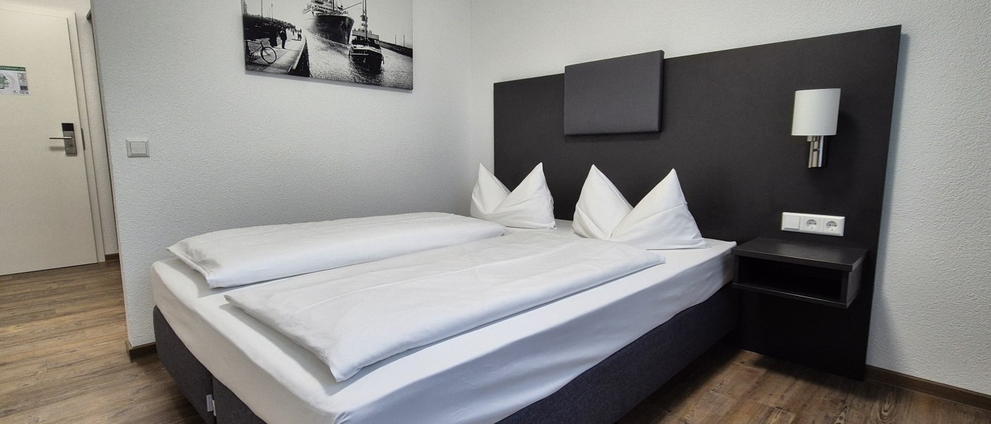 Modernes Hotelzimmer mit Doppelbett, wei&szlig;en Bettdecken, Nachttisch und Wandbild eines Schiffs. Helle W&auml;nde und Holzboden., &copy; MedientechnikKeuk