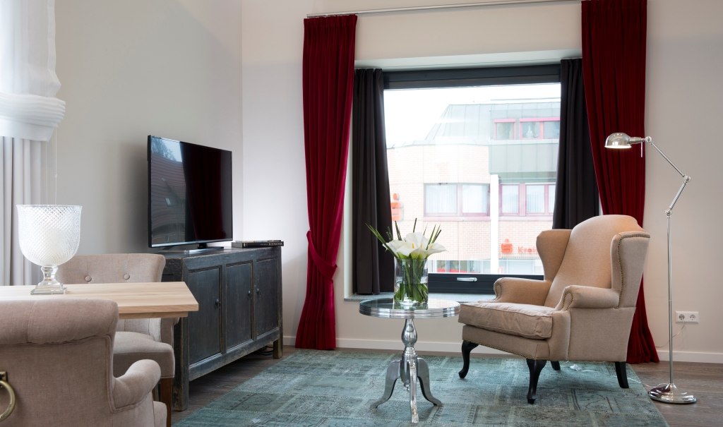 Elegant room with armchair, table, TV and large window with red curtains. The room is modern and inviting., © Hotel LOGIS Elegant room with armchair, table, TV and large window with red curtains. The room is modern and inviting., © Hotel LOGIS