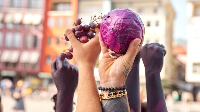 H&auml;nde halten roten Kohl und Trauben in die Luft. Die Arme sind teilweise bemalt. Im Hintergrund sind unscharfe Geb&auml;ude zu sehen., &copy; Theater Rampe