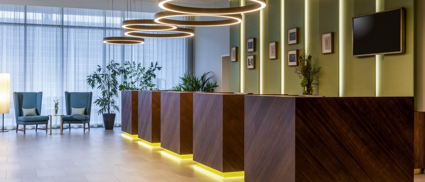 Moderne Rezeption mit Holztheken, hängenden Leuchtringen und grünen Pflanzen. Zwei blaue Sessel stehen links neben einem kleinen Tisch., © Mövenpick Hotel Stuttgart Messe & Congress