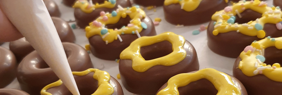 Chocolate rings are decorated with yellow icing and colorful sprinkles. One hand holds a piping bag., &copy; kesselglueck.de