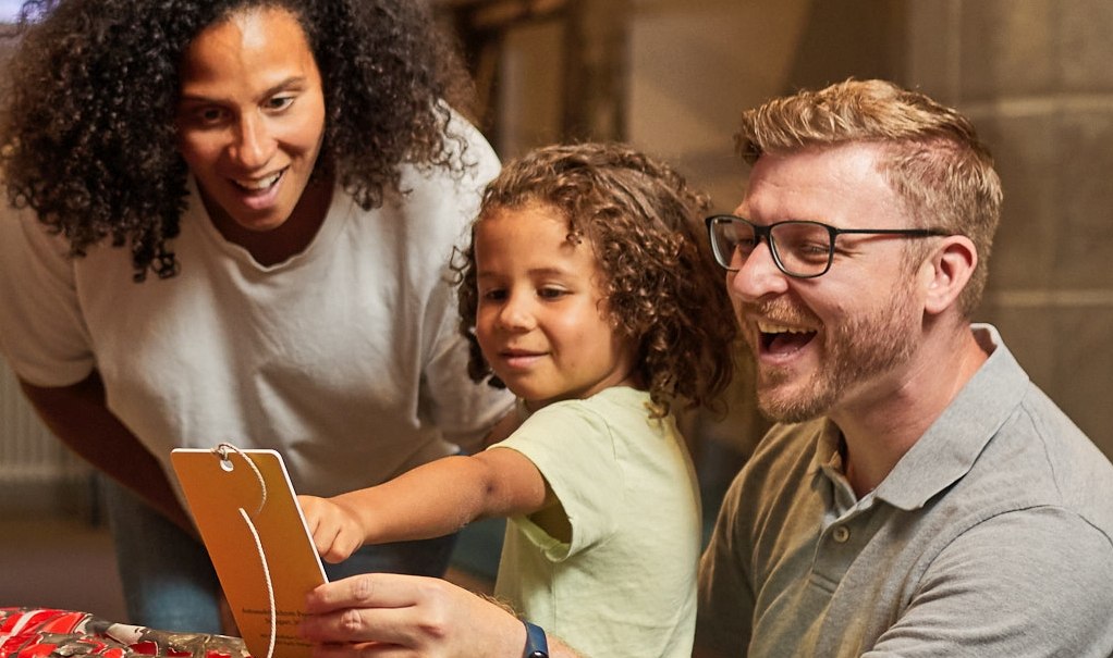 A child points to an object in a museum, accompanied by two smiling adults., © FTGRF.de A child points to an object in a museum, accompanied by two smiling adults., © FTGRF.de