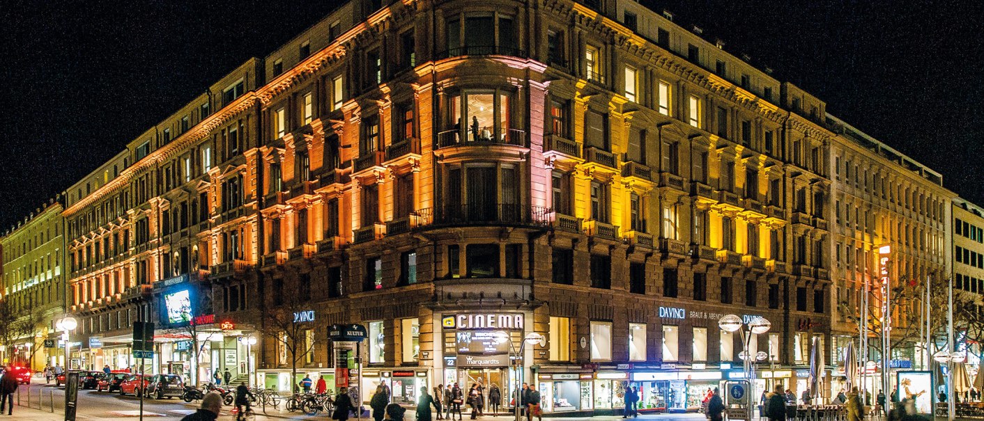 Ein großes, beleuchtetes Gebäude bei Nacht mit Geschäften im Erdgeschoss und Menschen auf der Straße., © Schauspielbühnen in Stuttgart