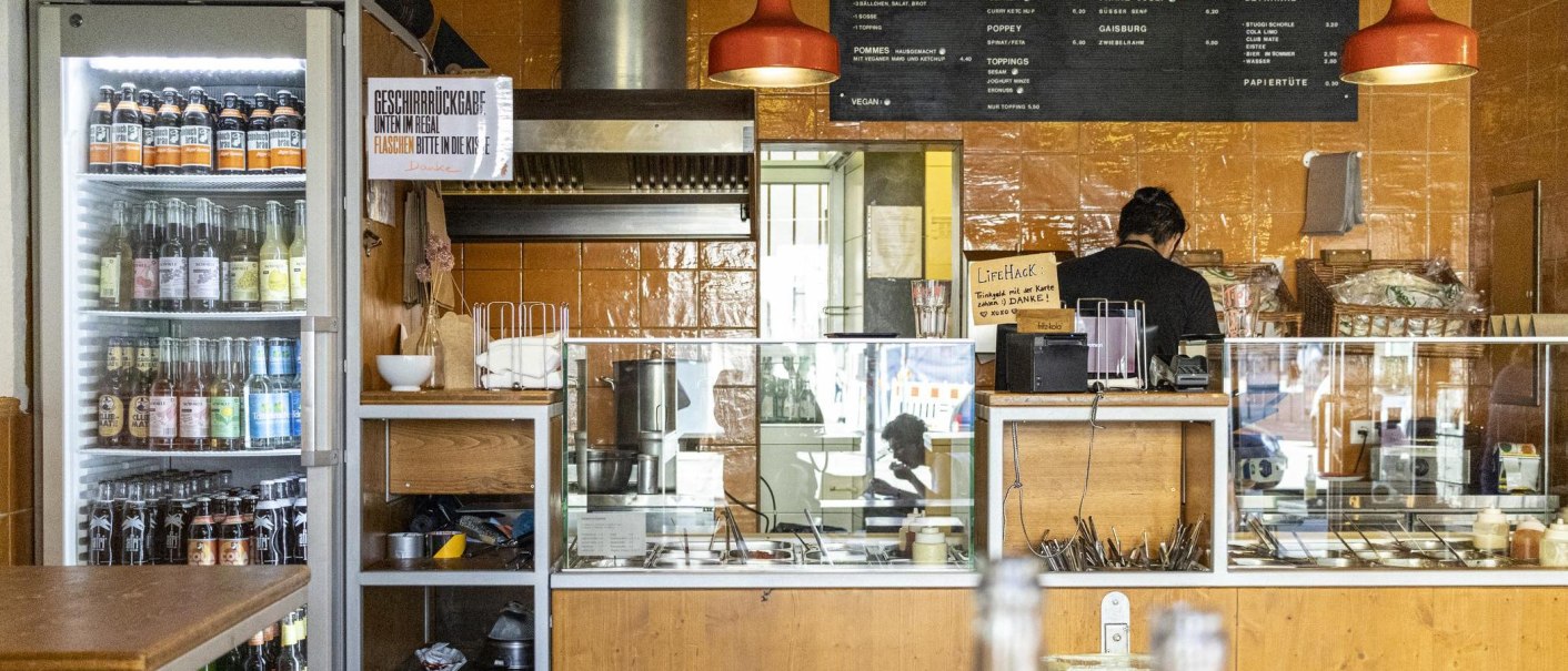 A snack bar with drinks fridge, counter and menu board. One person works behind the counter. The walls are tiled in orange., © SMG, Sarah Schmid