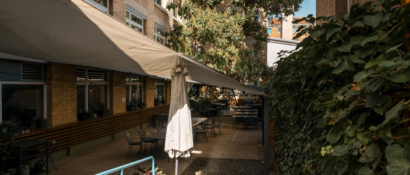 Innenhof des Künstlerhauses Stuttgart mit Sonnensegel, Tischen und Stühlen. Umgeben von Backsteinwänden und Pflanzen, bei sonnigem Wetter., © SMG, Sarah Schmid