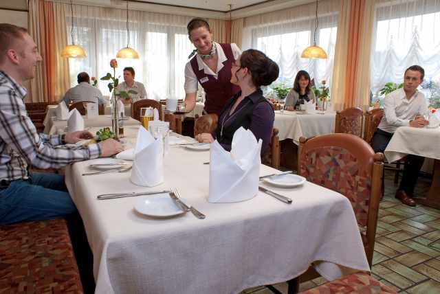 Ein Kellner bedient Gäste in einem gemütlichen Restaurant. Die Tische sind mit weißen Tischdecken und gefalteten Servietten gedeckt., © Ulli Kellenbenz