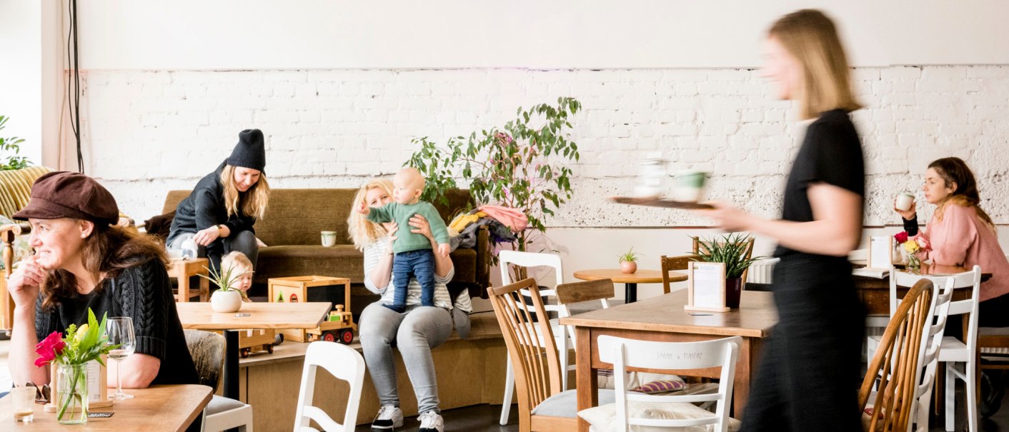 Gemütliches Café mit spielenden Kindern und Erwachsenen. Eine Bedienung serviert Getränke. Pflanzen und Holzmöbel schaffen eine einladende Atmosphäre., © Sven Weber Gemütliches Café mit spielenden Kindern und Erwachsenen. Eine Bedienung serviert Getränke. Pflanzen und Holzmöbel schaffen eine einladende Atmosphäre., © Sven Weber
