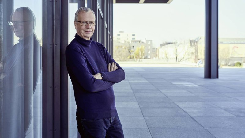 Ein Mann steht mit verschränkten Armen an einer Glaswand, seine Spiegelung ist sichtbar. Der Hintergrund zeigt eine moderne, urbane Umgebung., © Norman Posselt