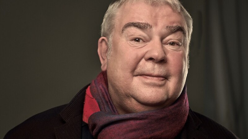 Ein Mann mit grauen Haaren tr&auml;gt einen roten Schal und l&auml;chelt in die Kamera. Der Hintergrund ist neutral gehalten., &copy; Theaterhaus Stuttgart e.V.