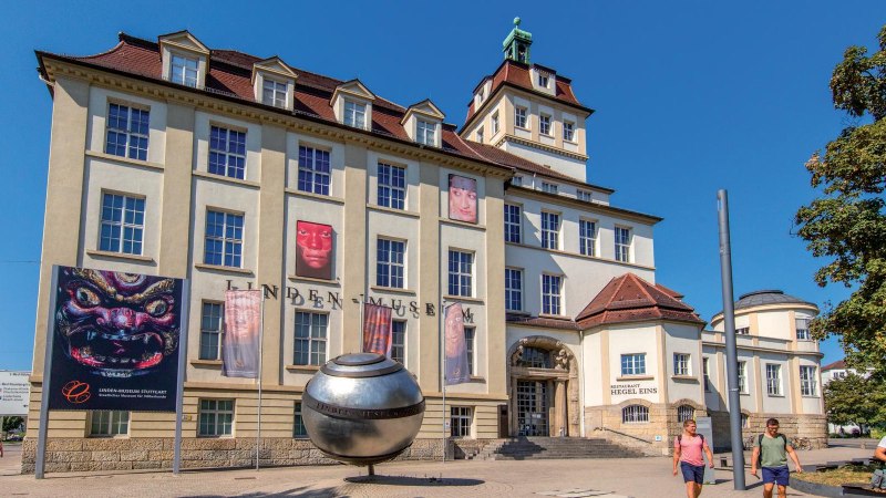 Das Linden-Museum in Stuttgart bei sonnigem Wetter. Vor dem Geb&auml;ude sind Plakate und eine gro&szlig;e silberne Kugel zu sehen. Menschen spazieren vorbei., &copy; Stuttgart-Marketing GmbH, Achim Mende