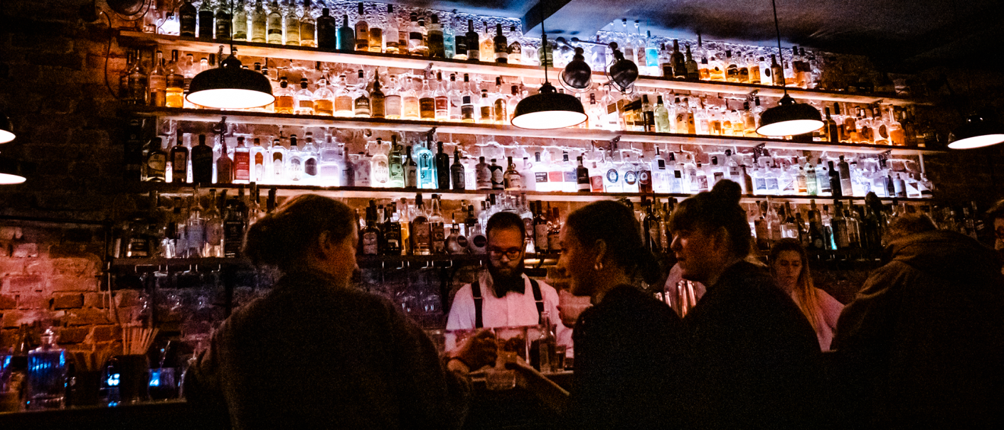 Eine stimmungsvolle Bar mit beleuchteten Flaschenregalen. Menschen unterhalten sich, während ein Barkeeper Getränke zubereitet., © Stuttgart-Marketing GmbH, Sarah Schmid