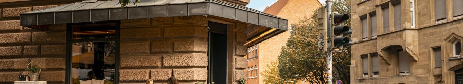 A street corner with a butcher's shop. Two people are sitting at a table in front of the store. A car drives past and there is a traffic light., &copy; SMG, Sarah Schmid