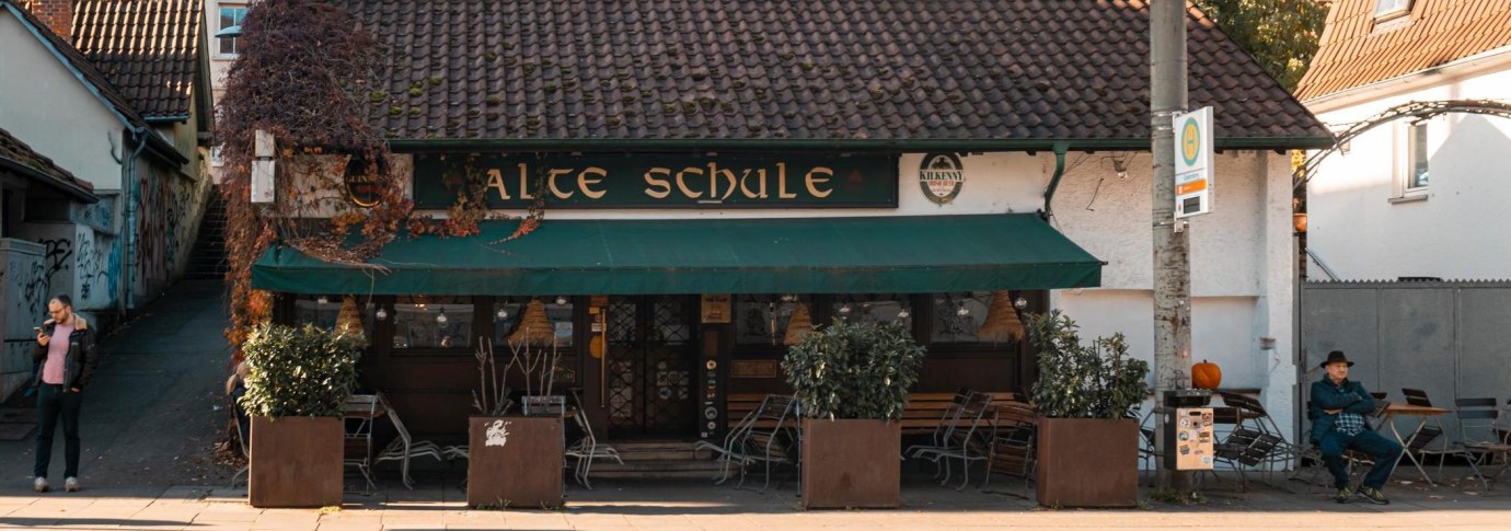 Ein gem&uuml;tliches Geb&auml;ude mit der Aufschrift 'Alte Schule', umgeben von Pflanzen. Ein Mann sitzt auf einer Bank, w&auml;hrend ein anderer sein Handy betrachtet., &copy; Stuttgart-Marketing GmbH, Sarah Schmid
