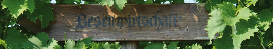 Ein Holzschild mit der Aufschrift 'Besenwirtschaft' ist von grünen Weinblättern umgeben., © SMG