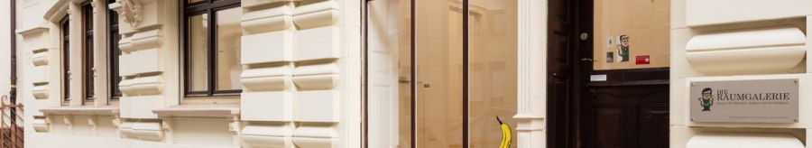 Eingang einer Galerie mit dem Schild 'Die Raumgalerie'. Eine Banane ist an der Glastür angebracht. Das Gebäude hat eine verzierte Fassade., © Dirk Wilhelmy Fotografie