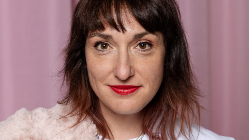 Nahaufnahme einer Frau mit braunen Haaren und rotem Lippenstift vor einem rosa Hintergrund. Sie tr&auml;gt ein wei&szlig;es Oberteil und einen flauschigen Schal., &copy; Julia Ruhland