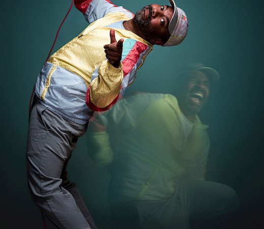 Ein Mann in bunter Jacke und Kappe posiert mit einem Mikrofon vor einem dunklen Hintergrund. Die dynamische Pose vermittelt Bewegung., &copy; Rosenau Kultur e.V.