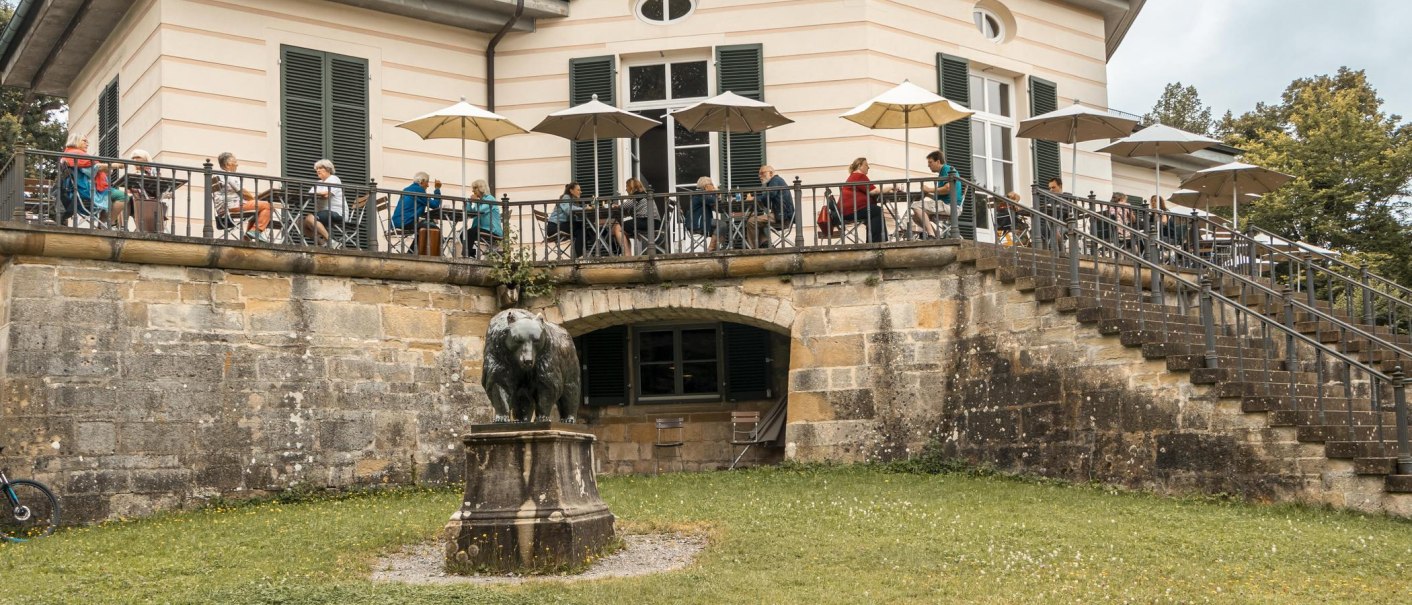Das Bärenschlössle mit Terrasse, auf der Menschen unter Sonnenschirmen sitzen. Eine Bärenstatue steht im Vordergrund auf einer Wiese., © SMG, Sarah Schmid