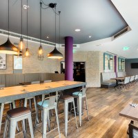 Modern breakfast area with long wooden tables, metal chairs and stylish hanging lamps. Stone walls and large windows create an inviting atmosphere., &copy; Premier Inn Germany