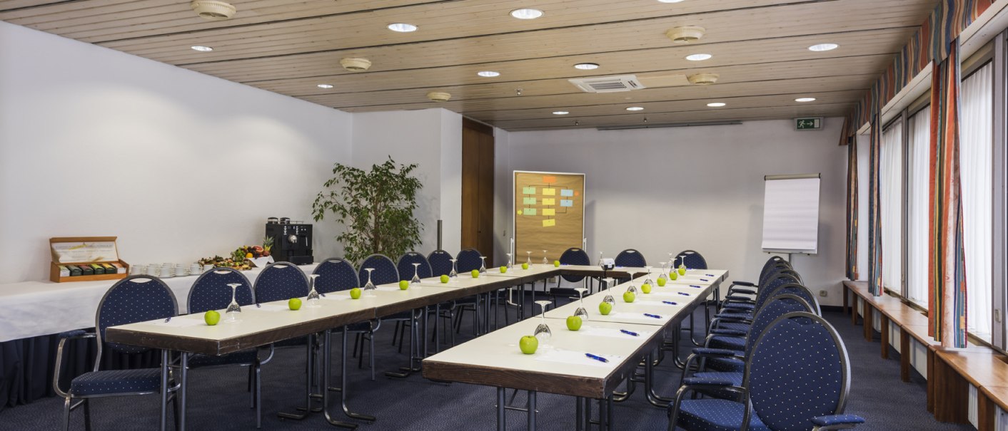 Conference room with a U-shaped table arrangement, blue chairs, green apples, and writing supplies. A flip chart and a bulletin board in the background., &copy; Hotel Berlin aZIS Hotel BetriebsGmbH
