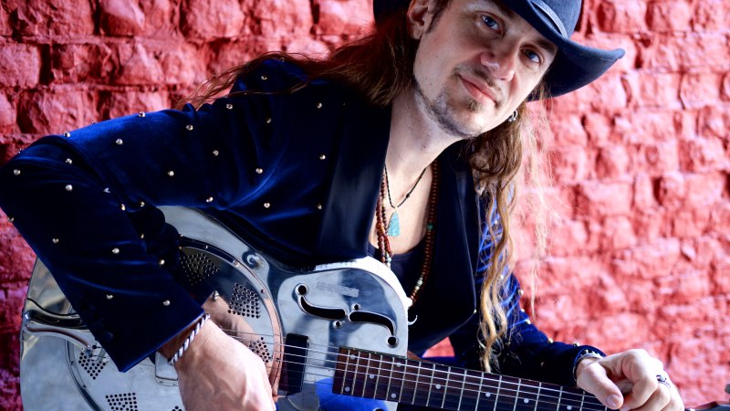 Person mit Cowboyhut spielt eine metallische Gitarre vor einer roten Backsteinwand. Sie trägt einen blauen Anzug mit Nieten., © Pressefoto Person mit Cowboyhut spielt eine metallische Gitarre vor einer roten Backsteinwand. Sie trägt einen blauen Anzug mit Nieten., © Pressefoto