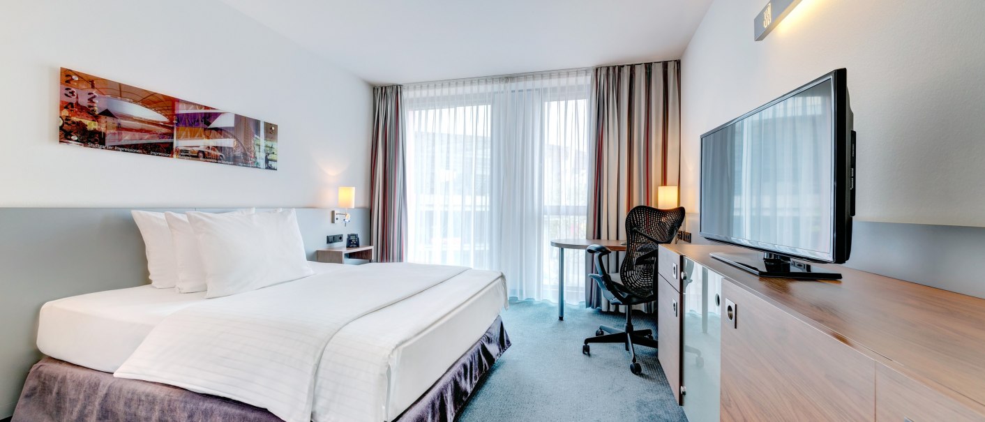 Modernes Hotelzimmer mit Queensize-Bett, Schreibtisch, Stuhl und Flachbildfernseher. Gro&szlig;e Fenster mit Vorh&auml;ngen lassen viel Licht herein., &copy; Hilton Garden Inn Stuttgart Neckarpark