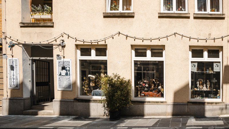 Fassade eines Antiquariats mit zwei großen Schaufenstern, dekoriert mit Antiquitäten. Eine Lichterkette hängt darüber, Pflanzen schmücken die Fenster., © SMG Stuttgart Marketing GmbH - Sarah Schmid Fassade eines Antiquariats mit zwei großen Schaufenstern, dekoriert mit Antiquitäten. Eine Lichterkette hängt darüber, Pflanzen schmücken die Fenster., © SMG Stuttgart Marketing GmbH - Sarah Schmid