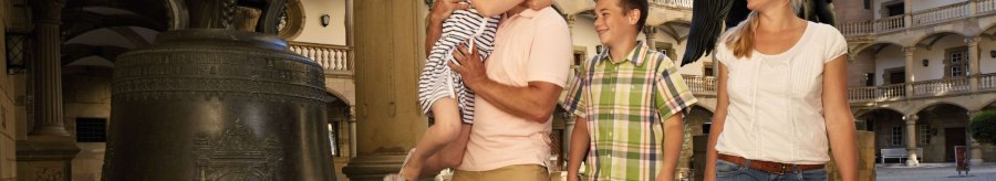 Eine Familie im Innenhof des Alten Schlosses Stuttgart. Ein Mann h&auml;lt ein Kind, w&auml;hrend eine Frau und ein Junge daneben stehen. Eine gro&szlig;e Glocke ist im Vordergrund., &copy; Stuttgart-Marketing GmbH Christoph D&uuml;pper