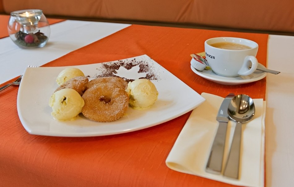 Plate with apple rings and vanilla ice cream, next to it a cup of coffee on an orange and white tablecloth. Cutlery lies on a napkin., © Hotel Neuwirtshaus Plate with apple rings and vanilla ice cream, next to it a cup of coffee on an orange and white tablecloth. Cutlery lies on a napkin., © Hotel Neuwirtshaus