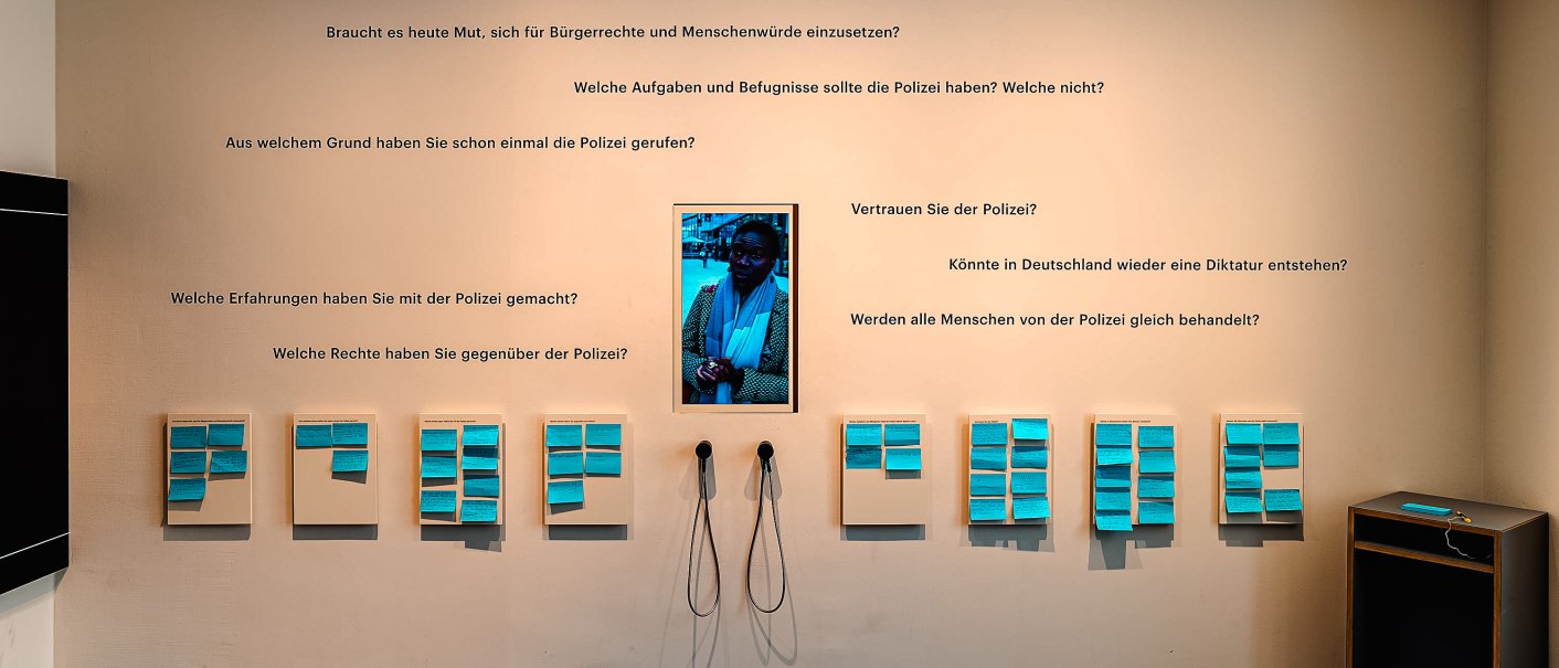 Ausstellungswand mit Fragen zur Polizei und Bürgerrechten, blauen Notizen und Kopfhörern. Ein Bildschirm zeigt eine Person. Beleuchtung von oben., © Daniel Stauch Photography